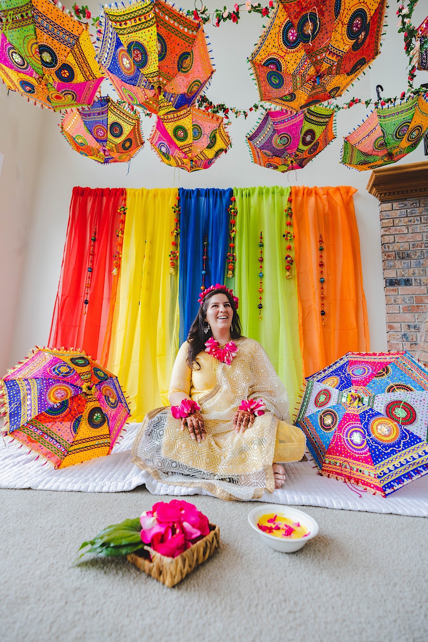 Bride to be sits in front of multicolored backdrop and colorful umbrellas before traditional Haldi celebration