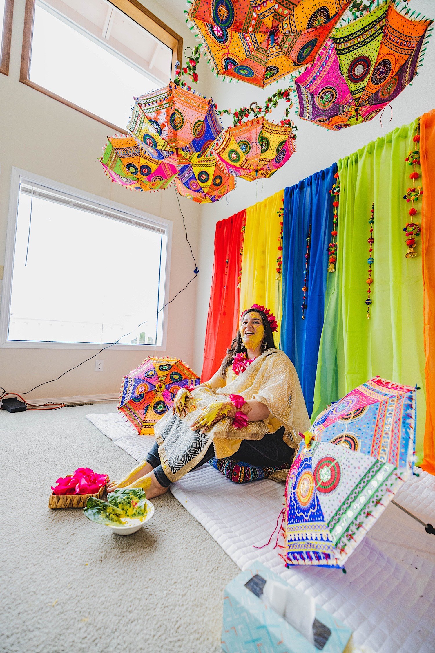 Bride to be covered in turmeric sits in front of multicolored backdrop and colorful umbrellas before traditional Haldi celebration