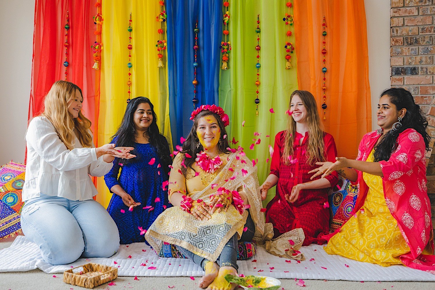 Bridesmaids throw flower petals at bride to be covered in turmeric at traditional Haldi celebration