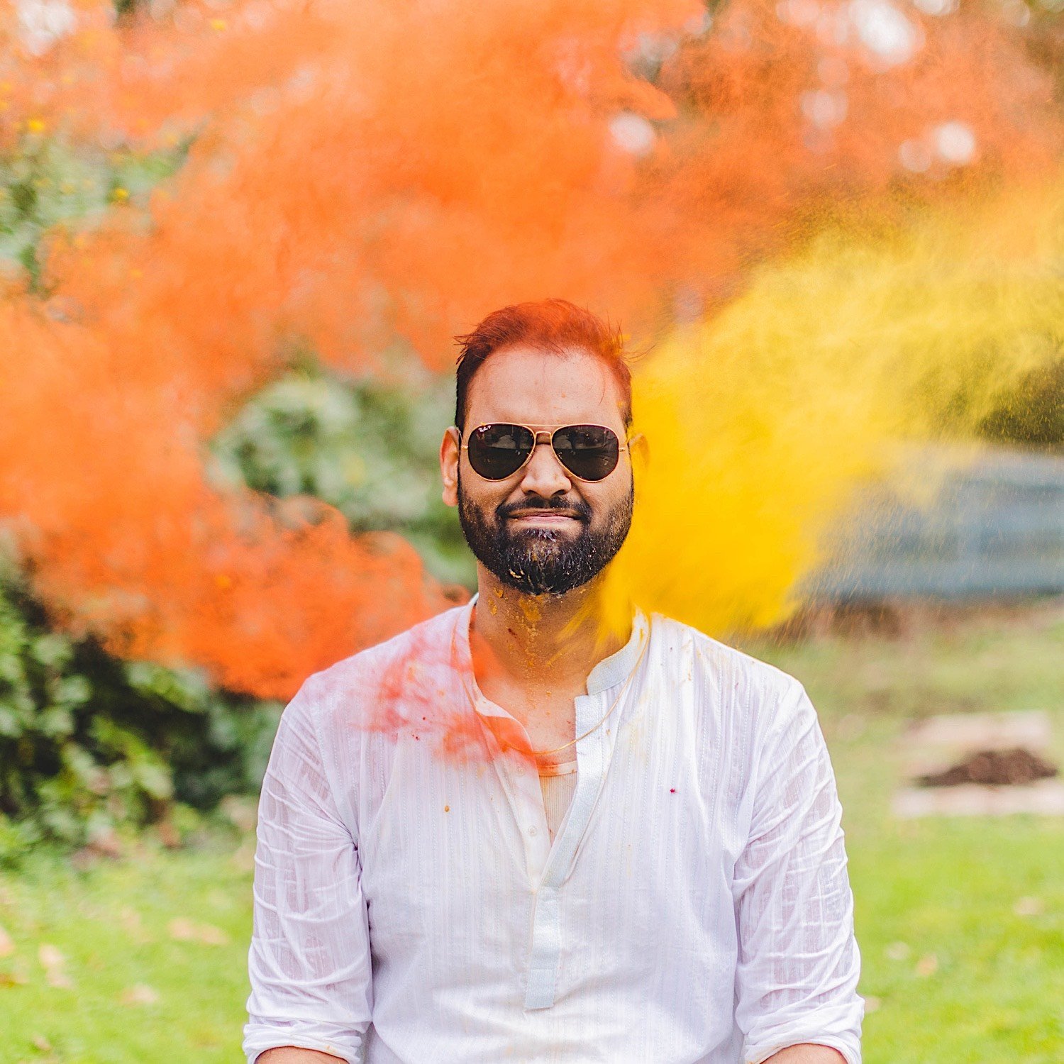 Groom wearing sunglasses has orange and yellow powder thrown at him during Haldi celebration