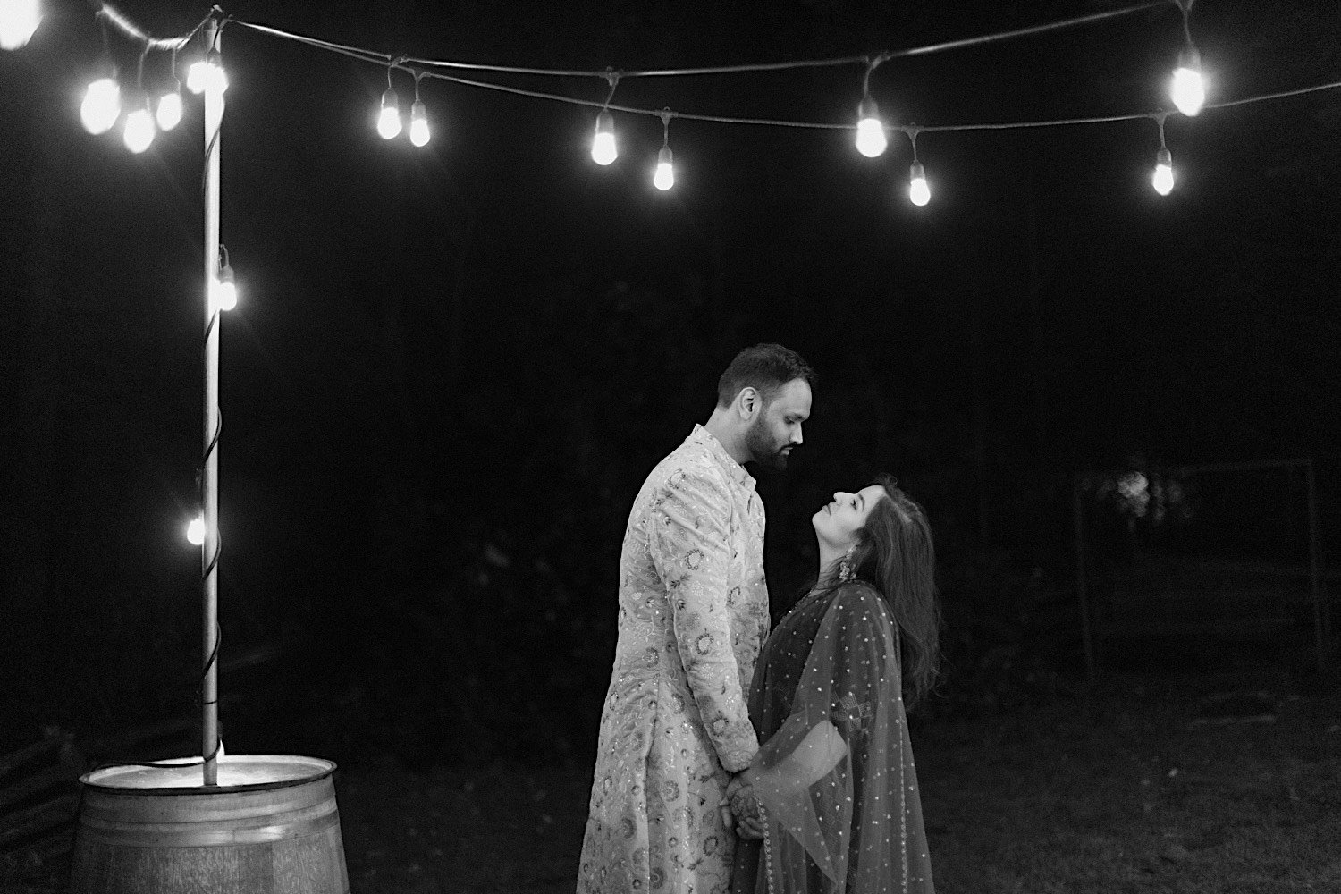Black and white photo of bride and groom standing underneath string lights looking at one another