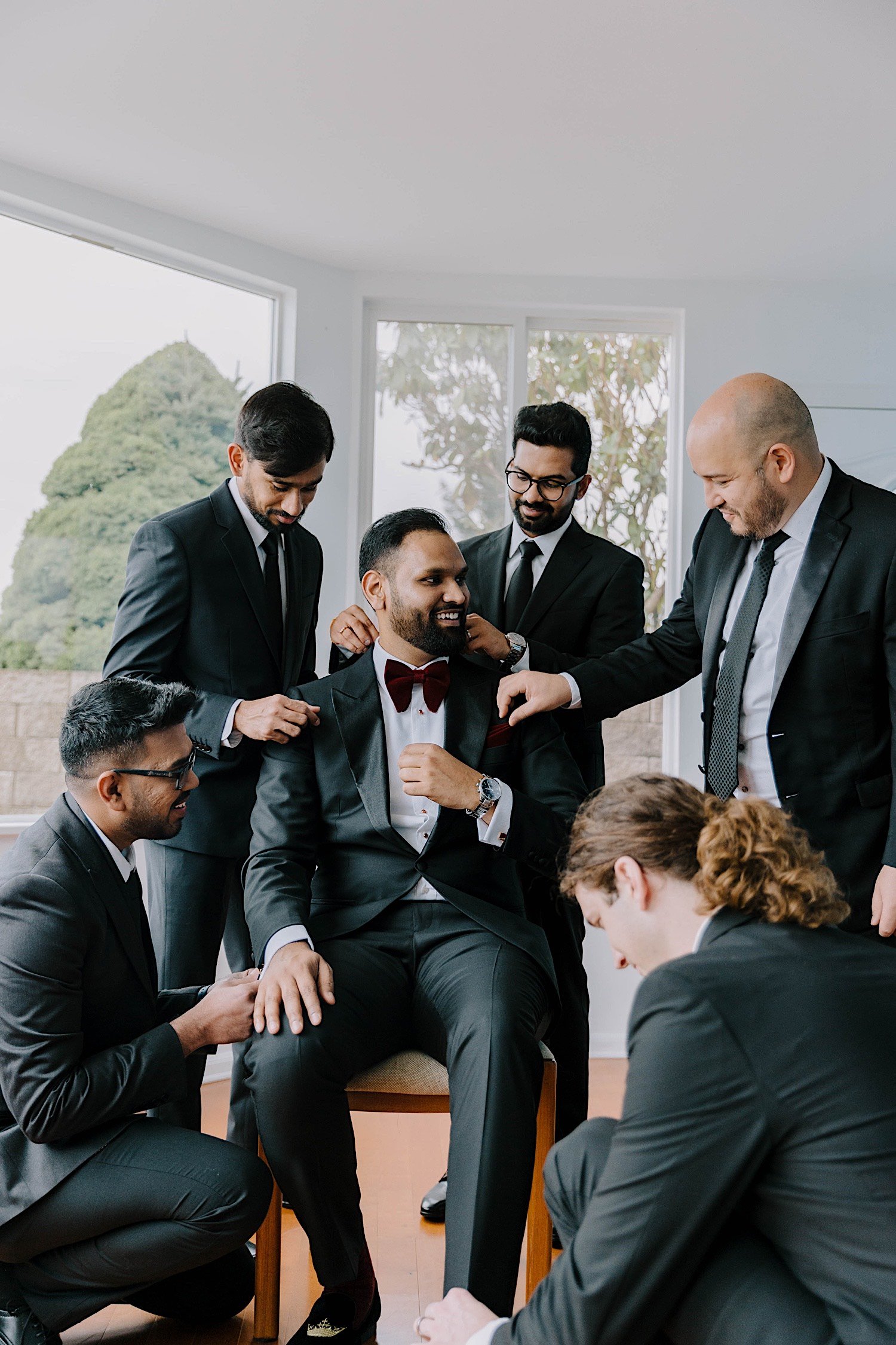 Groomsmen look over and touch up a seated groom as he laughs