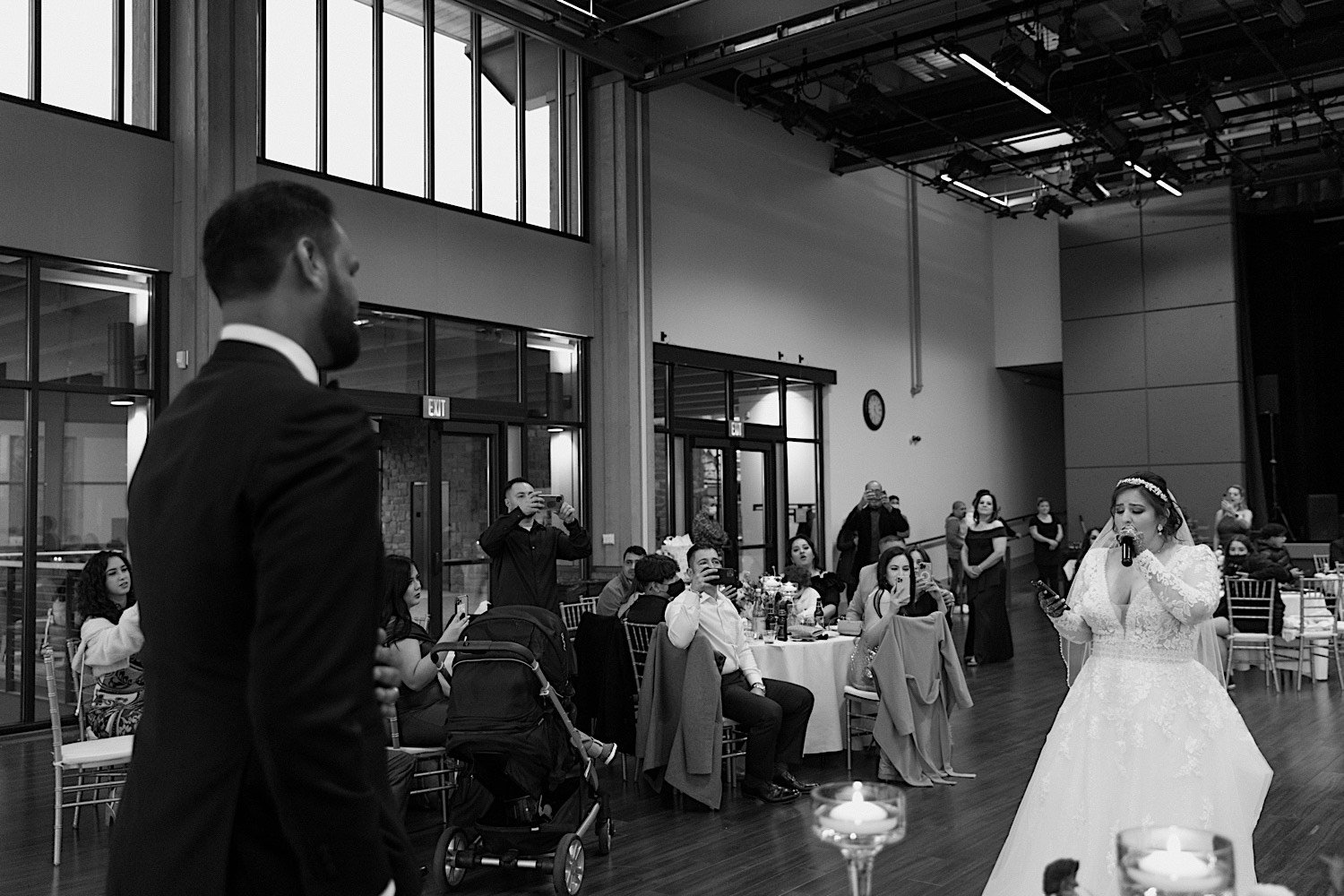 Black and white photo of bride singing to groom while guests watch and record in the background
