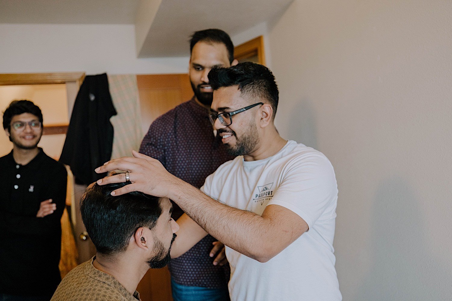 Man fixes grooms hair as they are getting ready for his wedding day with his two other friends smiling in the background