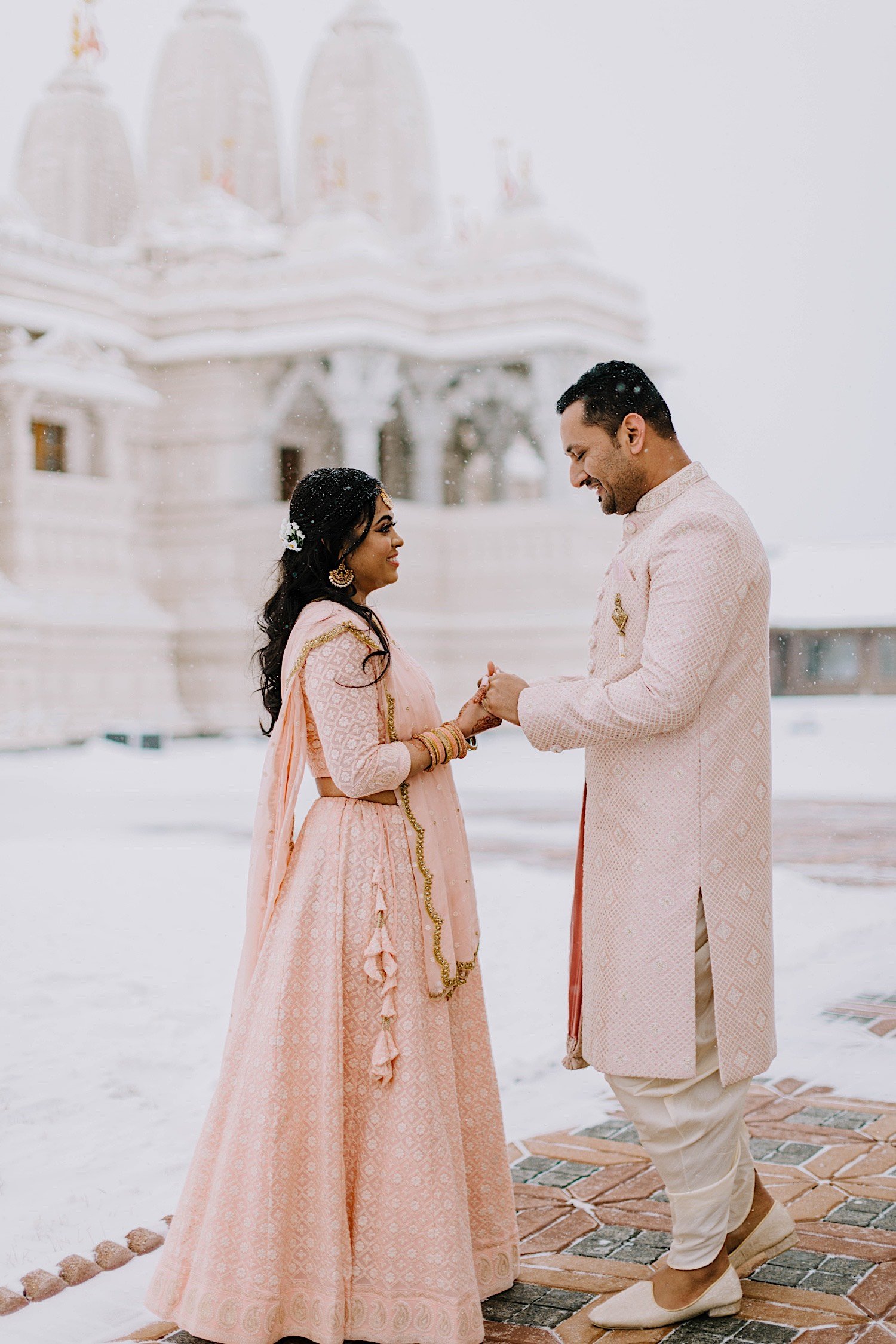Couple hold hands and smile at one another while wearing traditional Indian wedding attire with snow falling around them