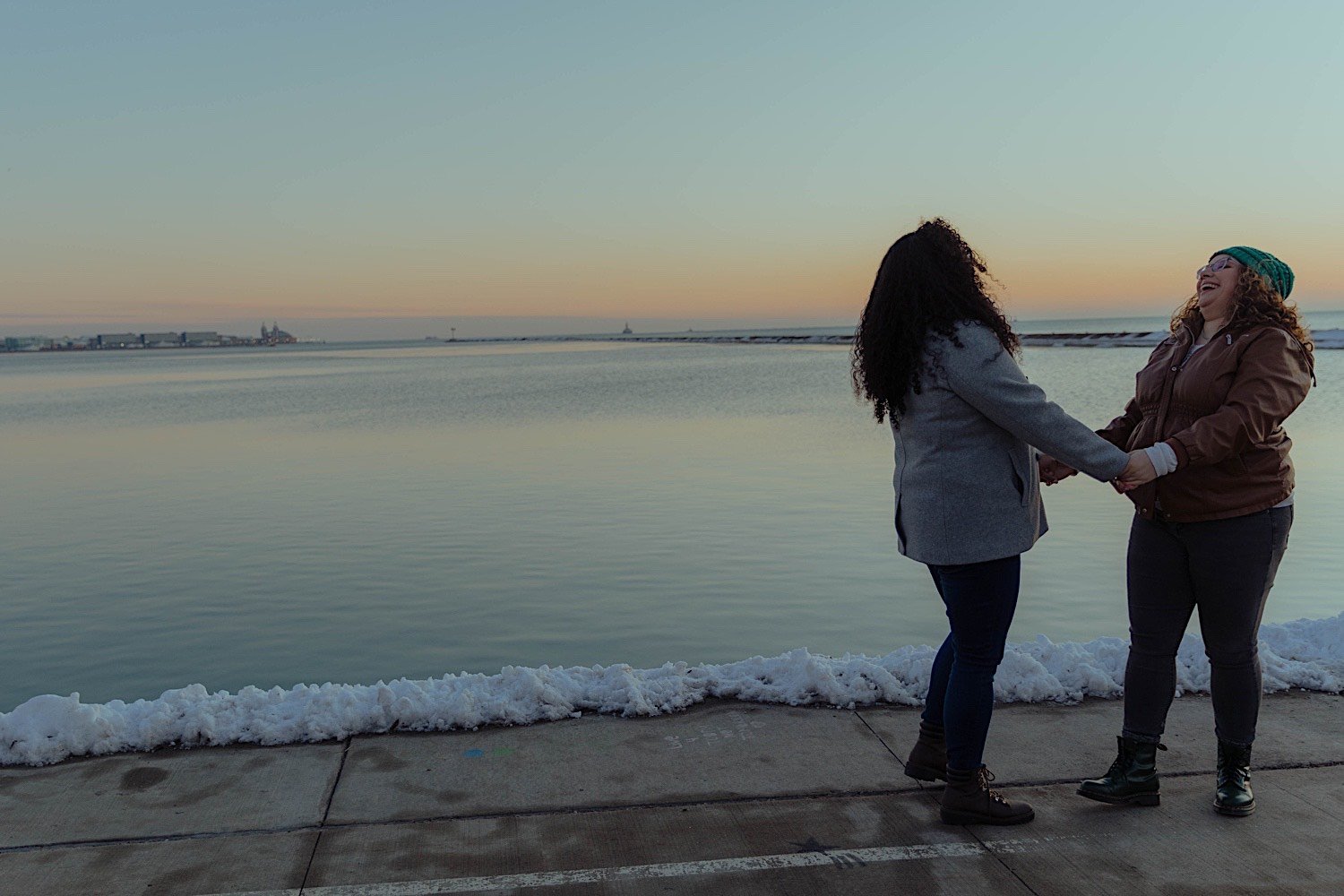 Couple laugh and hold hands during their museum campus engagement in front of a snowy Lake Michigan at sunrise