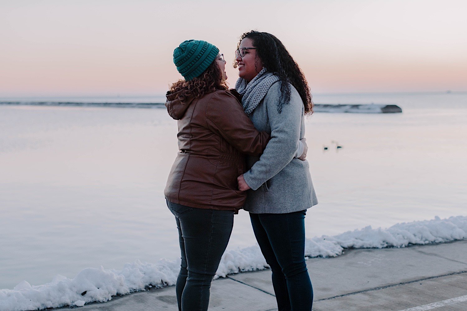 Couple embrace and smile at one another during their museum campus engagement in front of a snowy Lake Michigan at sunrise