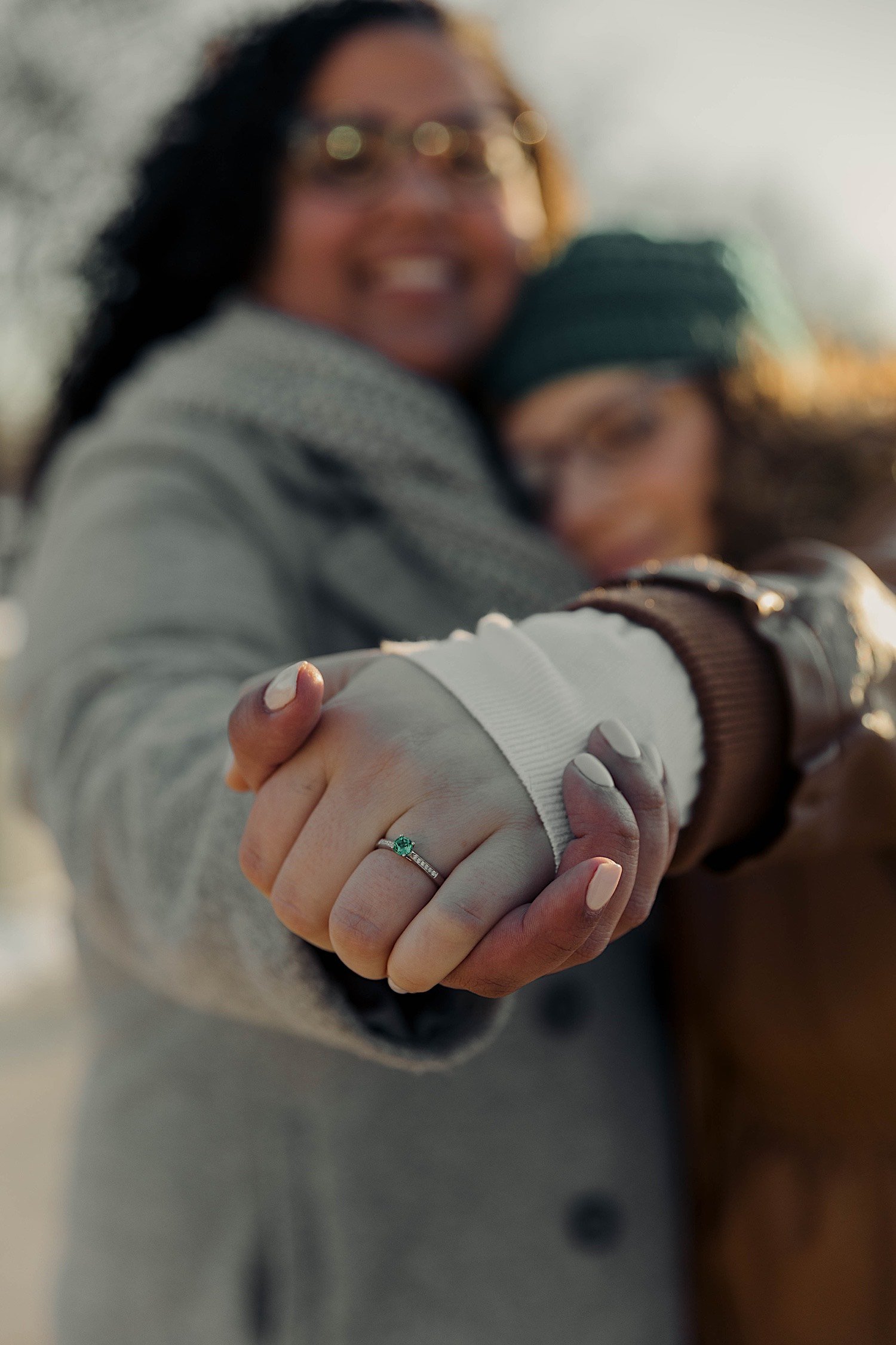 Couple hug and extend their hands towards the camera to show off engagement ring