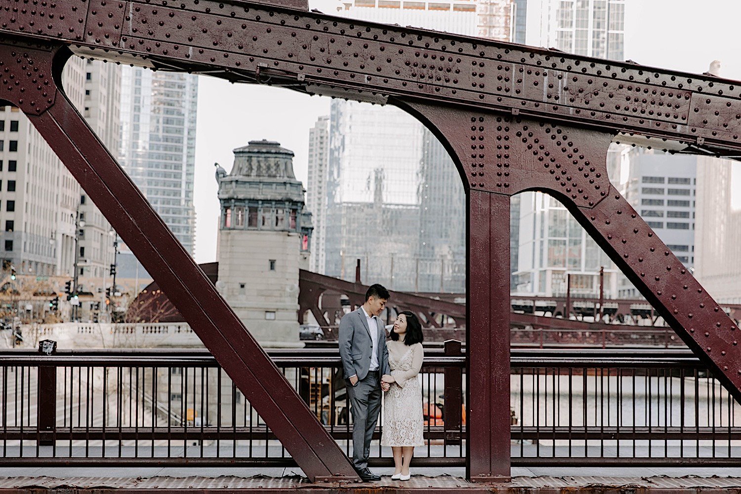 Couple stands next to each other and look at one another on a bridge in Chicago with the city in the background