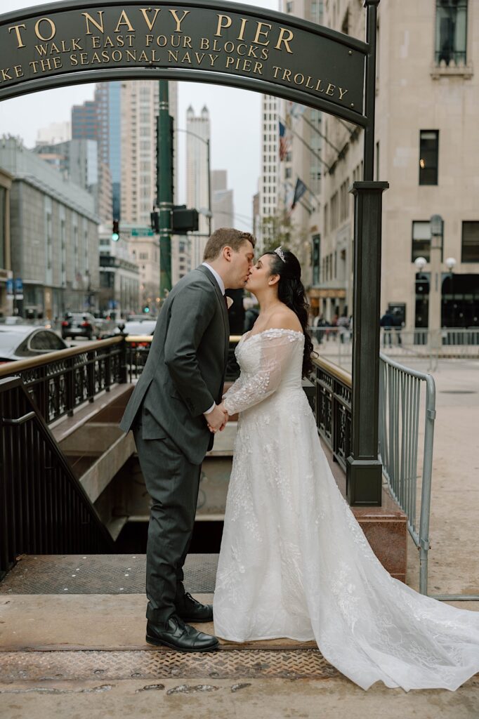 A bride and groom in their wedding gear kiss in front of a staircase leading down in the city  of Chicago. The sign above them reads " To Navy Pier"