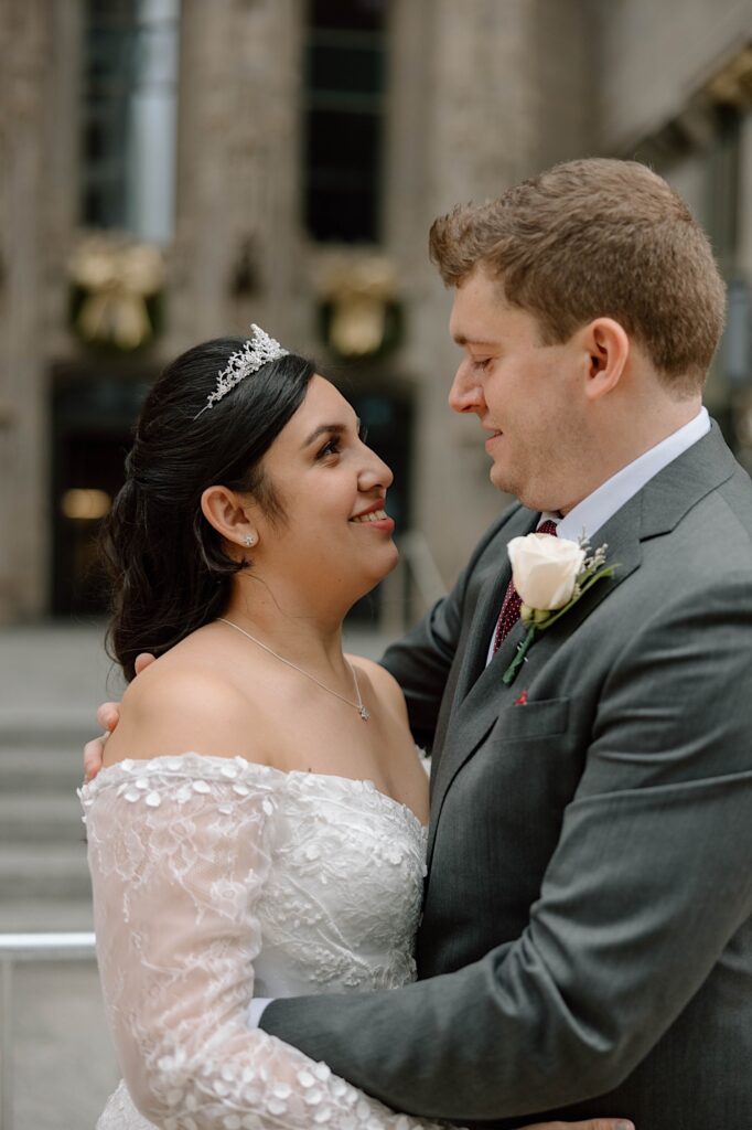 A bride and groom in their wedding attire embrace and look at one another while smiling. The background is a building out of focus.