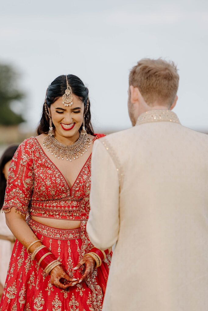 A bride wearing a red traditional Indian wedding dress and jewelry smiles while looking down. Her groom stands in front of her with his back to the camera.