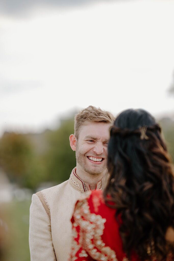A groom wearing a traditional Indian wedding suit smiles while looking at his bride in front of him. The bride is wearing a red dress and has her back to the camera.