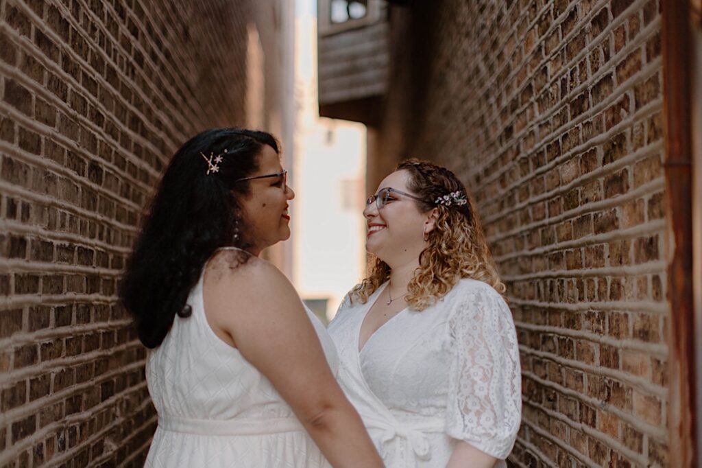 Two brides stand together in a brick alleyway smiling at one another in their wedding dresses.