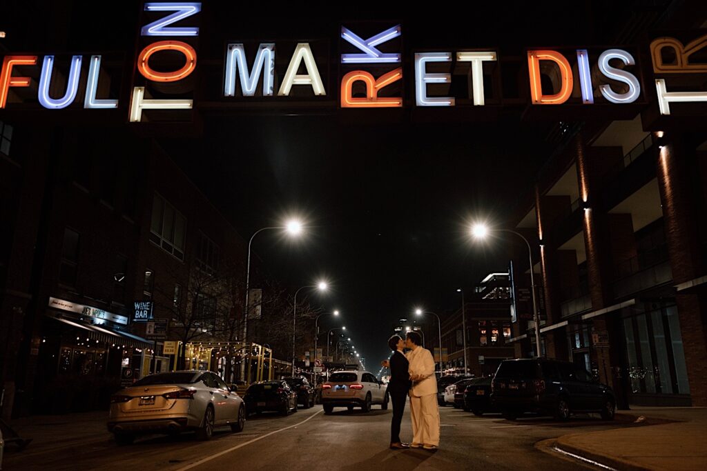 A newlywed couple kiss in the middle of a Chicago street at night outside of their intimate wedding venue. The colorful sign above them reads "Fulton Market District"