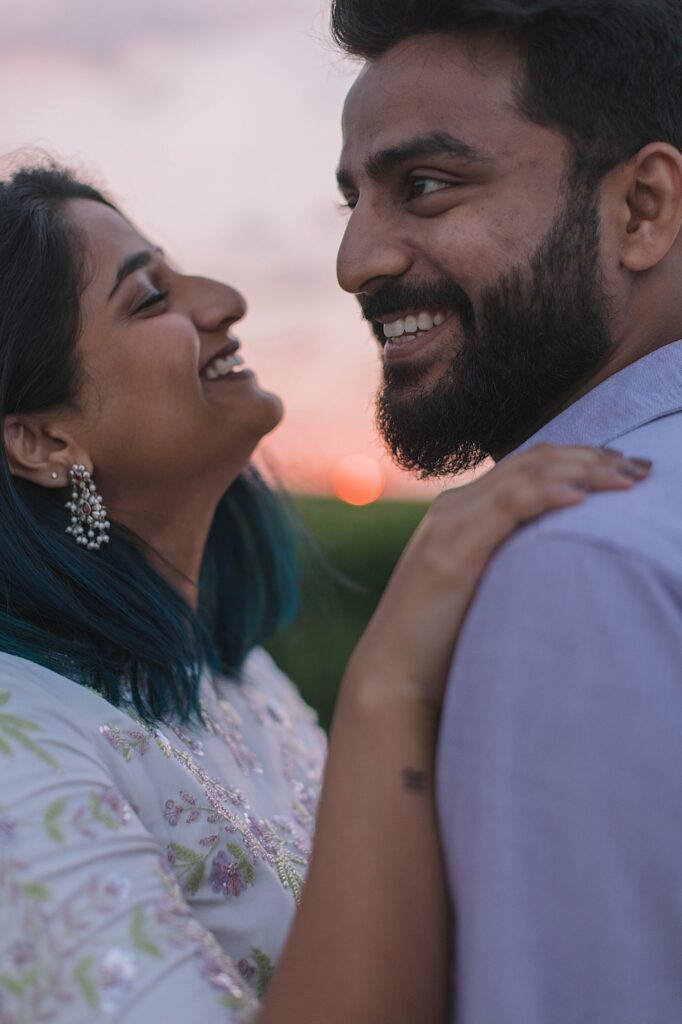 A man and woman embrace and smile while the sun sets behind them.