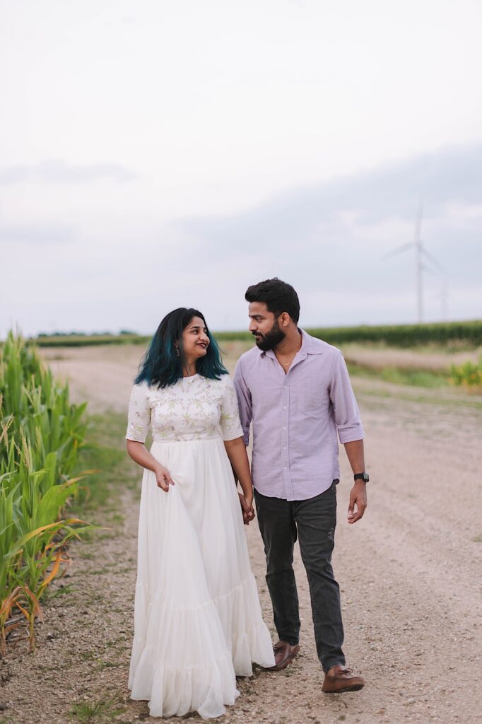 A couple walk hand in hand down a dirt road while looking at one another, in the background is a wind turbine and cornfields.