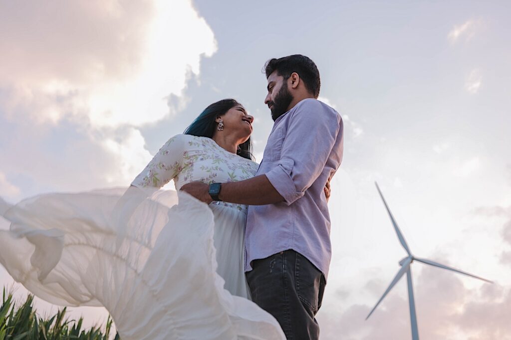 A couple embrace and dance during their newlywed session in a Chicagoland cornfield. Behind them is the sky, a wind turbine and some corn.