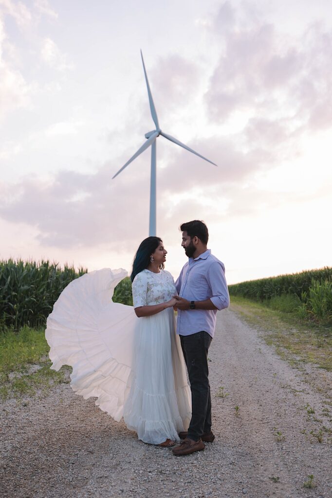 A couple embrace and look at one another on a dirt road surrounded by corn and with a wind turbine behind them