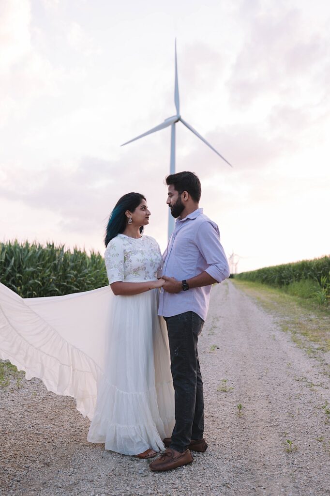 A couple embrace and look at one another on a dirt road surrounded by corn and with a wind turbine behind them