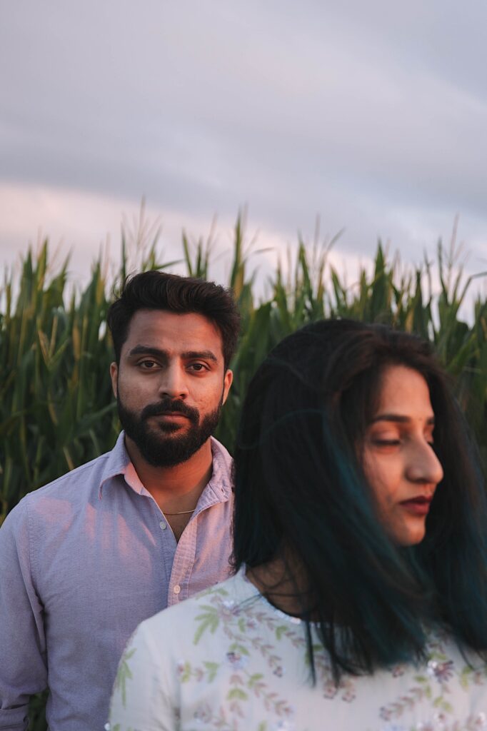 A man looks at the camera while his wife stands in front of him and has her eyes closed, behind them is a cornfield.