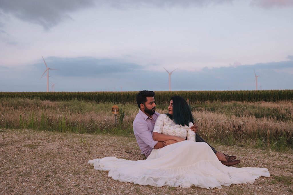 A couple sit on a dirt road and look at one another during their newlywed session in a Chicagoland cornfield, behind them are fields of corn and 3 wind turbines.