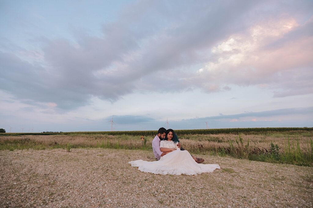 A couple sit on a dirt road and look at the ground during their newlywed session in a Chicagoland cornfield, behind them are fields of corn and 3 wind turbines.