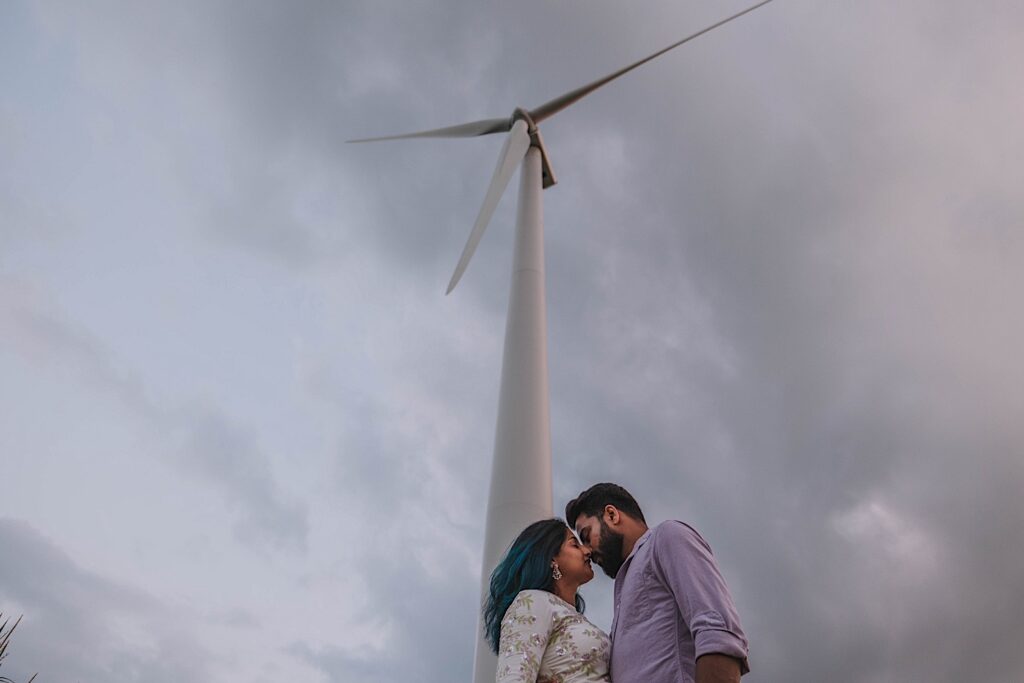 A couple that are about to kiss stand underneath a wind turbine during their newlywed session in a Chicagoland corrnfield.