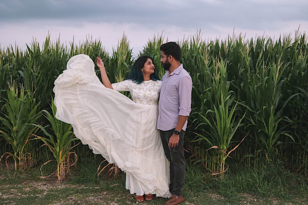 A couple stand next to one another and look at each other during their newlywed session in a Chicagoland cornfield. The woman is flinging her dress in the air and there are rows of corn behind them.
