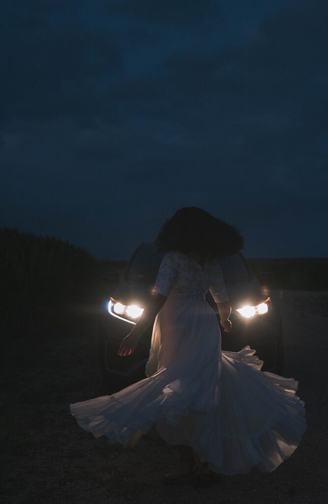 A woman twirls her dress at night while illuminated by the car behind her.