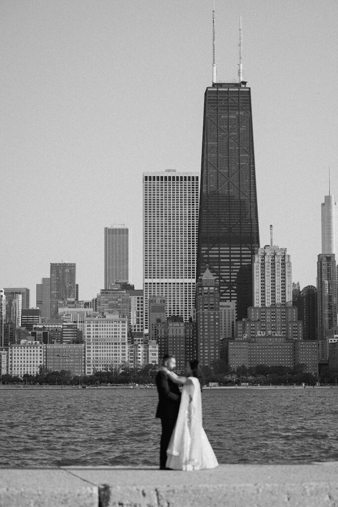 Black and white photo of a couple in their wedding attire embracing in front of Lake Michigan and the Chicago skyline. The couple is out of focus while the background is in focus