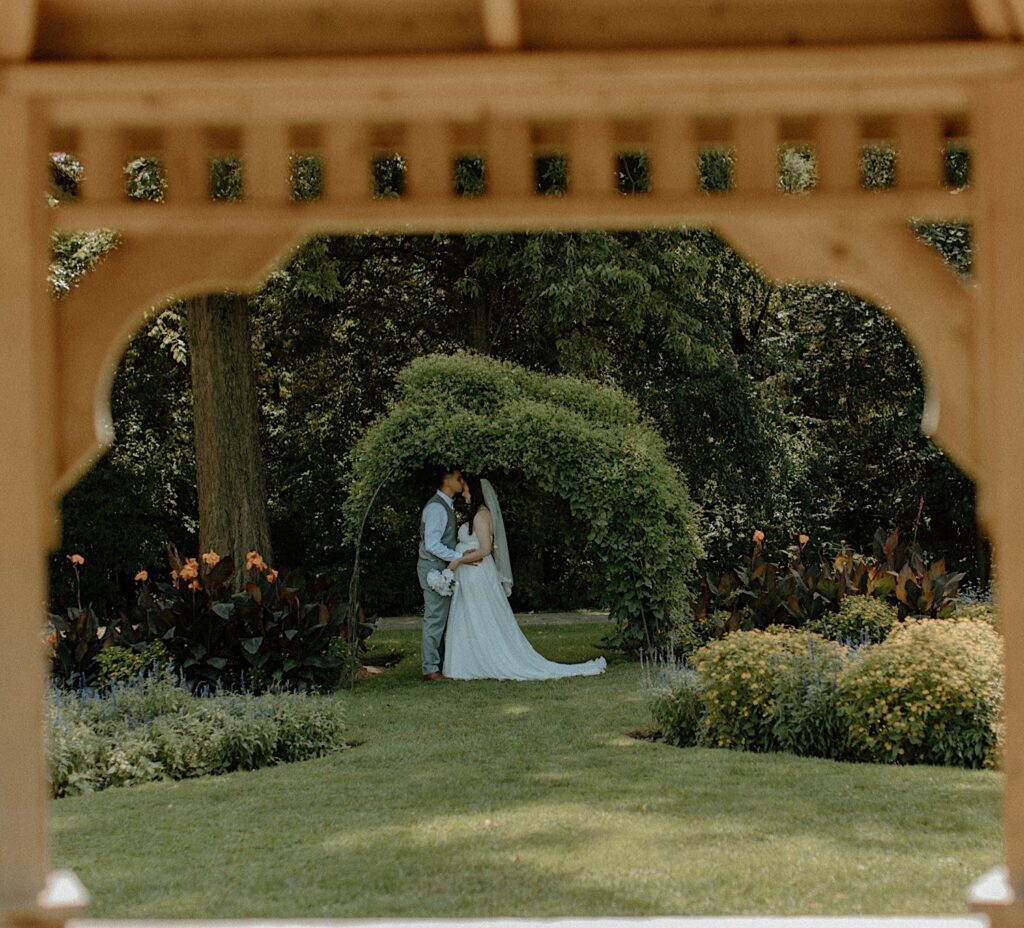 A couple kiss in a garden underneath an archway of leaves while in their wedding attire